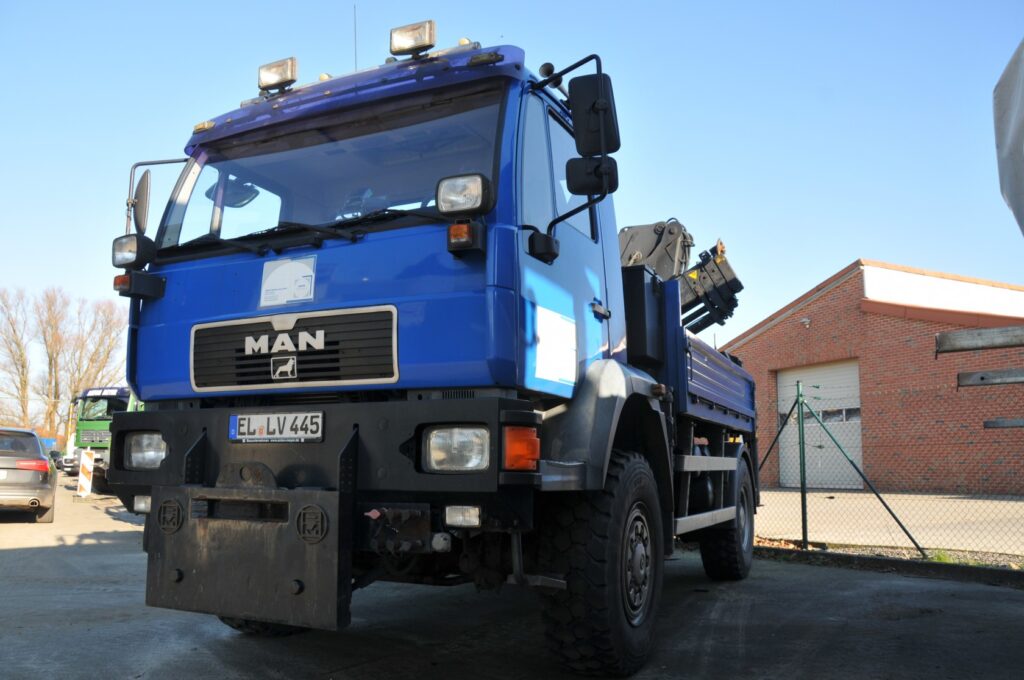 Blauer Lkw mit Kranausrüstung auf einem Hof, Gebäude mit rotem Klinker im Hintergrund.